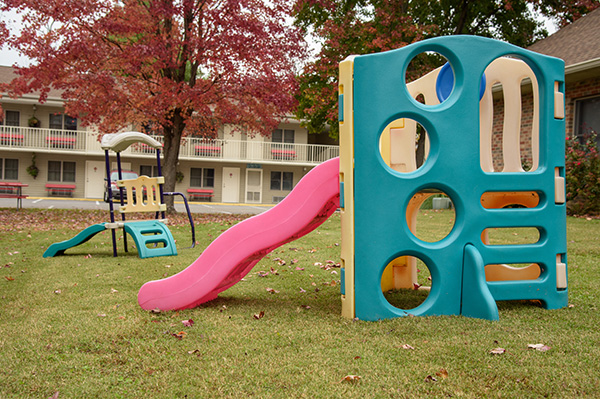 A playground with a blue and pink play structure and a tree with red leaves in front of a hotel building.