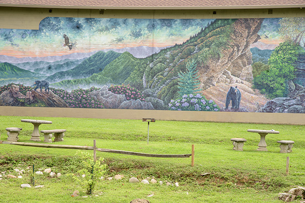 A large mural depicting a mountain range and wildlife is on the side of a building, with benches and a fence in the foreground.